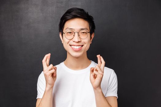 Close-up portrait of hopeful young anticipating asian guy praying, cross fingers good luck and smiling excited as making wish, want dream come true, standing black background pleading.
