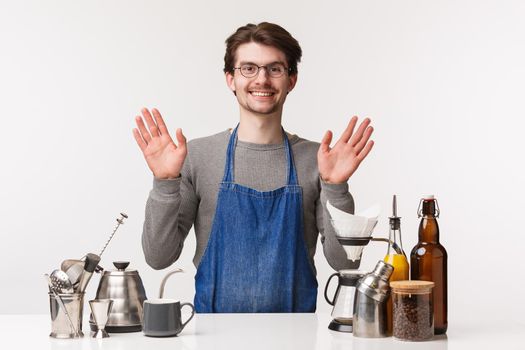 Barista, cafe worker and bartender concept. Portrait of friendly outgoing man in apron, invite clients have drink in his cafe, making coffee, saying hi waving in greeting hello sign, white background.