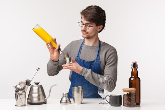 Barista, cafe worker and bartender concept. Portrait of serious-looking professional male employee in apron near bar counter pouring orange juice in shaker, making drink or coffee.