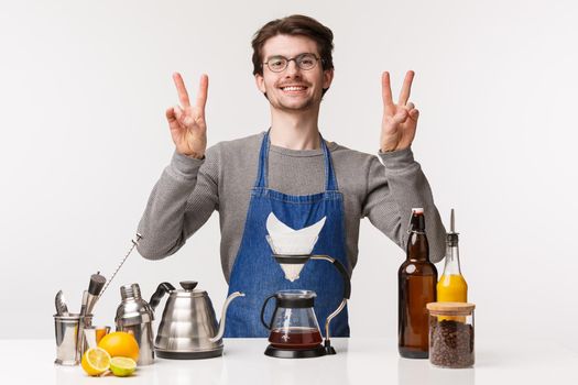 Barista, cafe worker and bartender concept. Portrait of cheerful friendly young male employee informal greeting dear customers, show peace signs and smiling made filter coffee.