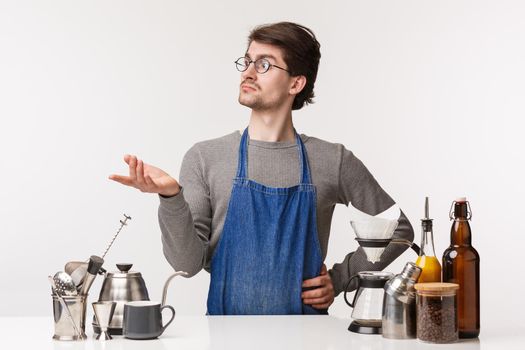 Barista, cafe worker and bartender concept. Confused young serious-looking guy working in restaurant, cafeteria, standing in apron near bar counter with coffee equipment, look away puzzled.