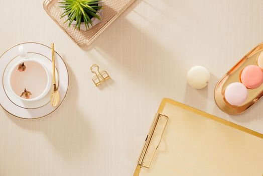 Modern gold stylized home office desk with folder, macaroons, coffee mug on beige background. Flat lay, top view lifestyle concept.`