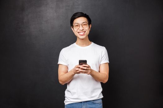 Technology, gadgets and people concept. Friendly smiling young asian guy with mobile phone, texting friends or using social media app, order taxi in application, look satisfied, black background.