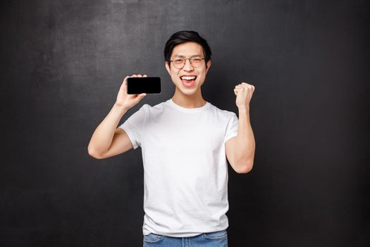 Portrait of lucky excited asian guy winning money on sport bet, fist pump and celebrating, showing his best score in mobile game on display, hold smartphone and triumphing, black background.