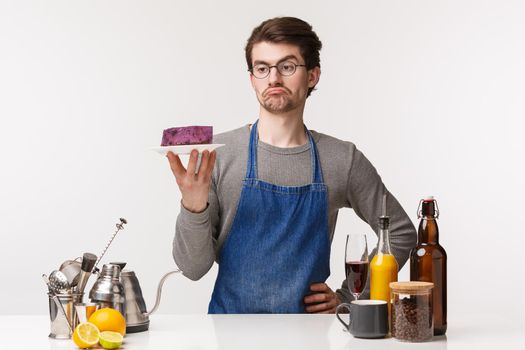 Barista, cafe worker and bartender concept. Portrait of young male employee cooked dessert, looking at cake on plate with skeptical uncertain expression, standing bar counter.