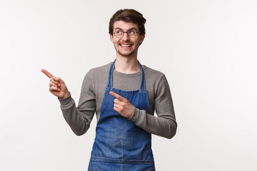 Cheerful funny young man in apron, barista enjoying good day at coffee shop, look and pointing upper left corner pleased, found great new chemex equipment, white background.