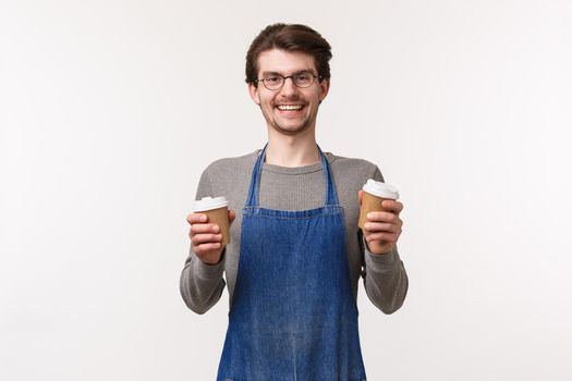 Portrait of outgoing carefree, smiling friendly male employee in apron, barista made delicious coffee for customers, hold two take-away cups and laughing, inviting try best cappuccino in town.
