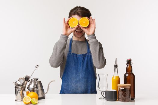 Barista, cafe worker and bartender concept. Portrait of cheerful happy male employee in blue apron making drink, holding two slices of orange like eyes mask and smiling camera, stand near bar counter.