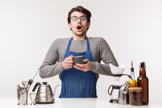 Barista, cafe worker and bartender concept. Shocked startled young male employee in apron, gasping astonished holding tea cup and staring amazed being scared while prepare coffee, white background.