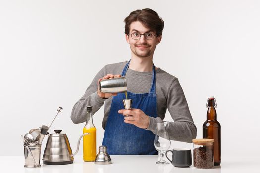 Barista, cafe worker and bartender concept. Portrait of enthusiastic happy young male employee in apron, making coffee, pouring drink in shaker, mixing flavours, standing white background.