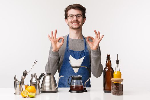 Barista, cafe worker and bartender concept. Portrait of cute friendly smiling caucasian male employee in apron guarantee you will like coffee, make filter with kettle, show ok sign.