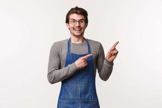 Excited, happy smiling caucasian male employee at coffee shop, barista or bartender in apron grinning enthusiastic stand white background, pointing upper right corner at promo.