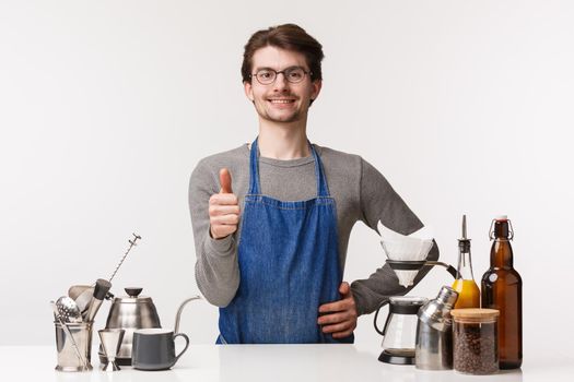 Barista, cafe worker and bartender concept. Portrait of pleased good-looking young man employee in apron, show thumb-up sign and smiling confident, guarantee you will like coffee, approve.