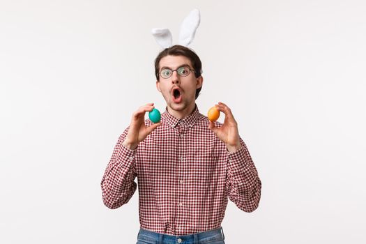 Traditions, religious holidays, celebration concept. Funny joyful young man with beard in glasses, wear cute rabbit ears and hold two painted eggs, playing game on Easter day, white background.