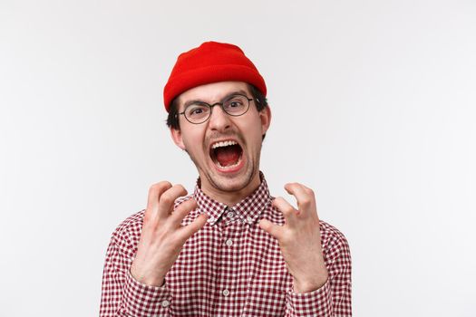 Close-up portrait of distressed and angry young funny hipster guy with moustache wear red beanie, glasses, squeez hands annoyed and bothered, screaming from pissed-off feeling, white background.
