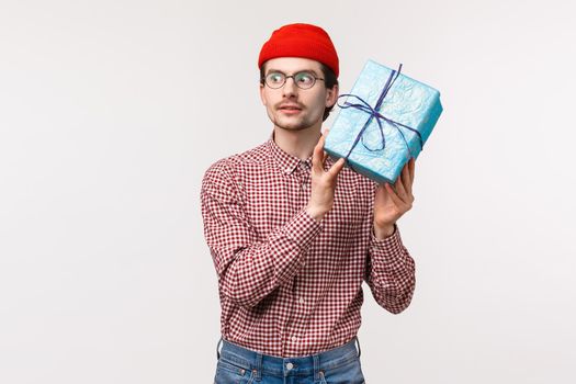 Waist-up portrait excited caucasian bearded guy in red beanie, shaking blue wrapped gift box near ear as trying guess whats inside, look thrilled, unboxing present on b-day party celebration.