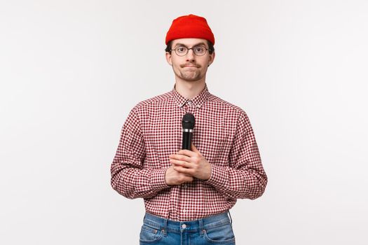 Close-up portrait of shy and geeky cute young bearded guy in glasses and red beanie, looking at tv screen and holding micrphone, getting ready to sing karaoke, standing white background.