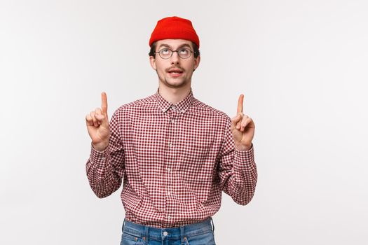 Waist-up portrait funny creative young man with mousatche in glasses and red beanie, pointing fingers up and rolling eyes to top as seeing something cool, standing white background.