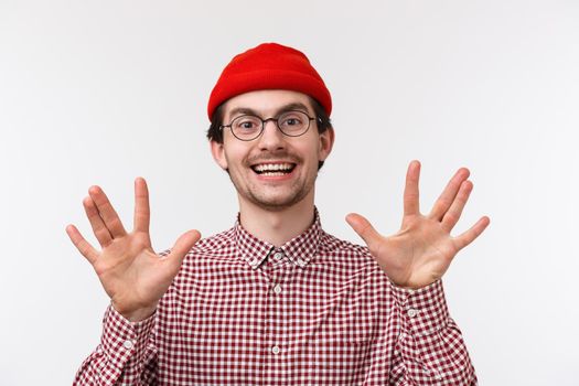 Close-up portrait funny hipster guy in red beanie and glasses make spok gesture showing fingers and smiling, discuss favorite tv series with friends geeks, standing white background upbeat.