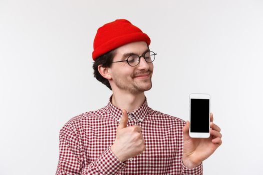 Technology and people concept. Close-up portrait pleased happy young hipster man in red beanie and glasses, looking satisfied at mobile phone display, make thumb-up approve smartphone gadget.