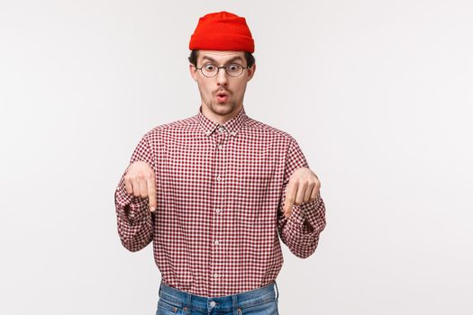 Wow incredible you see this awesome thing. Impressed and speechless good-looking caucasian man in red beanie and glasses, folding lips astonished and staring pointing down, white background.