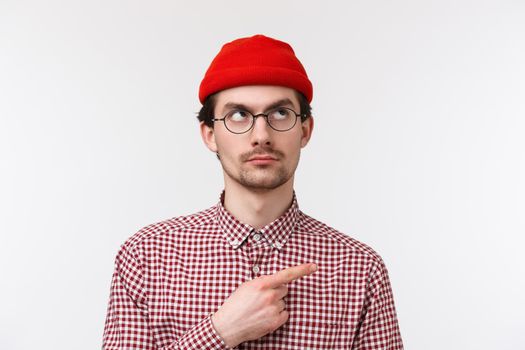 Skeptical good-looking young hipster guy in red beanie and check shirt, glasses, peeking doubtful, looking pointing upper right corner with serious expression, standing white background.