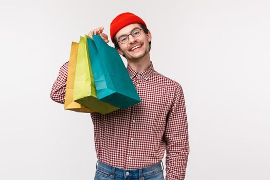Treat yourself day. Pleased and happy smiling caucasian guy likes shopping, holding bags and look camera satisfied as bragging with his new staff he bought in store, white background.