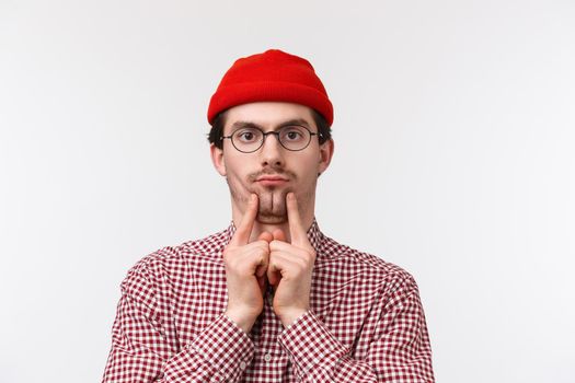 Funny and cute bearded caucasian guy in glasses and red beanie, squeez cheeks to make double-chin mimicking plump boss at work as if copying him mocking, standing white background.