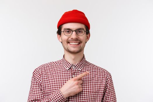 Close-up portrait handsome friendly-looking hipster male in red beanie and glasses, smiling satisfied and happy, pointing finger upper right corner, showing cool product or place hang out.