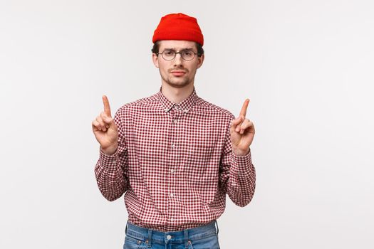 Waist-up portrait skeptical and unamused bored young man with beard, wear glasses and red beanie, look reluctant camera, pointing fingers up at something uninteresting with disappointment.