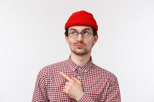 Close-up portrait skeptical and doubtful handsome bearded guy in red beanie, checked shirt and glasses, smirk unsatisfied and hesitant pointing upper left corner at something bad and unimpressive.
