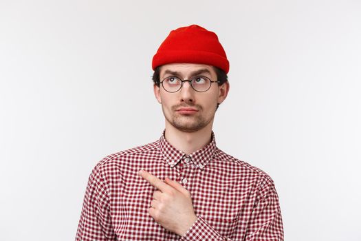 Skeptical and unimpressed handsome young hipster guy look judgemental and displeased, pointing staring upper left corner disappointed, standing white background reluctant. Copy space
