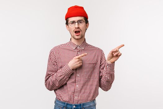 Waist-up portrait unimpressed reluctant and bored young man in glasses and red beanie yawning uninterested as pointing upper right corner at something that not worth attention, white background.