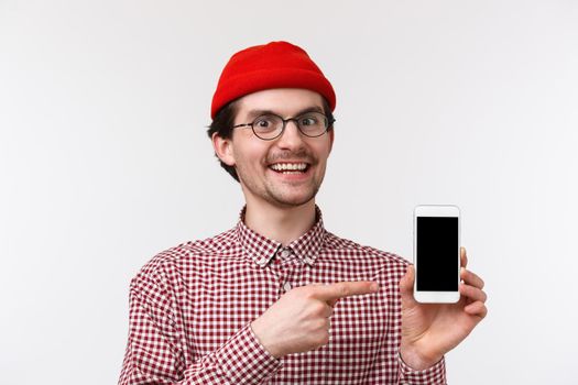 Technology and people concept. Close-up portrait excited pleased young man in red beanie and glasses, recommend subscribe to streaming service or buy app, point mobile phone display, smiling.