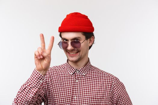 Close-up portrait funny and outgoing hipster man in red beanie, sunglasses and check shirt, getting ready summer vacation, showing peace gesture and smiling, stand white background.