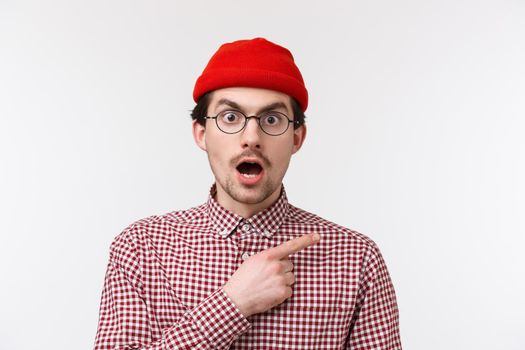 Close-up portrait surprised and shocked young caucasian guy with moustache, wear glasses and red beanie, drop jaw and stare camera as if hear shocking news, pointing upper right corner.