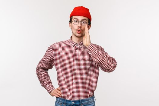Waist-up portrait of funny young guy with beard and moustache in red beanie, glasses, calling someone, searching for friend in crowd, hold hands near mouth gossiping, whisper secret.