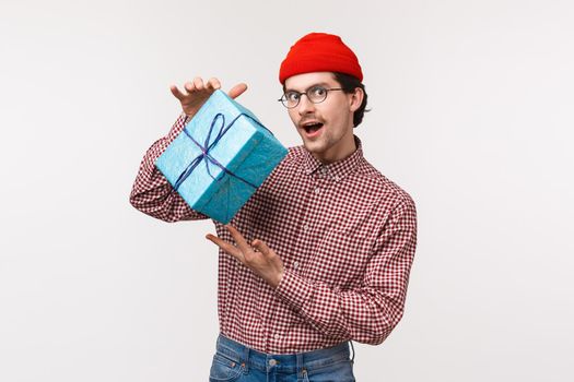Guy trying make intrigue as prepared present for girlfriend, shaking wrapped box with gift, wonder whats inside, look mysterious and cunning camera, wear red beanie checked shirt, white background.