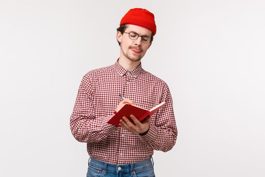 Waist-up portrait creative hipster guy in red beanie, glasses, stick tongue thoughtful while writing note in notebook, making list, help solve equation for younger brother, stand white background.