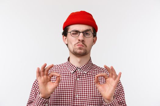 Close-up portrait impressed good-looking young hipster guy in red beanie, glasses, smirk pleased, show not bat okay gesture satisfied with good result, standing white background delighted.