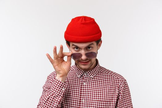 Close-up portrait of suspicious funny young guy in red beanie and checked shirt, take-off sunglasses and look from under forehead as gossiping, hinting or telling secret, spying on someone.