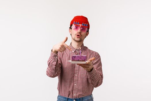 Celebration, holidays and lifestyle concept. Funny cheerful male hipster friend having fun at b-day party, wear glasses and pointing at birthday cake asking to blow out candle, white background.