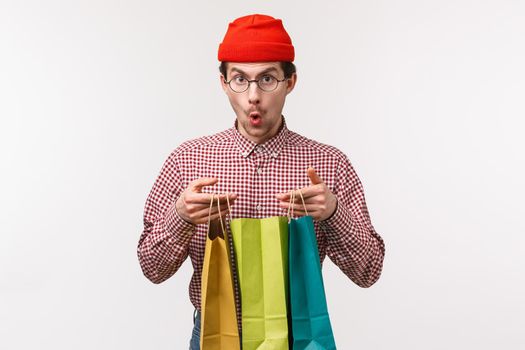 Waist-up portrait excited and surprised handsome young man with moustache, wear glasses and beanie, looking amazed camera say wow as open shopping bags with gifts, stand white background.