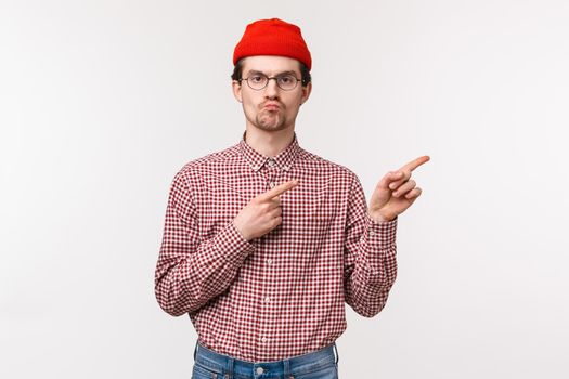 Skeptical serious-looking bothered young geeky man in red beanie and glasses, pouting with unsatisfied grimace pointing upper right corner, annoyed seeing something awful, express dislike.
