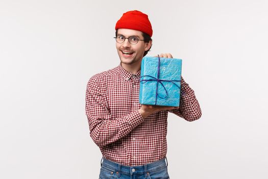 Waist-up portrait friendly funny cute man bought present for his mate, holding wrapped gift and smiling, buy surprise gift as going to b-day party, standing white background in red beanie.