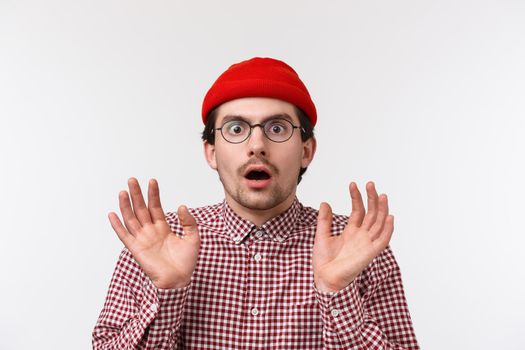 Speechless and concerned young shocked caucasian guy found out shocking news, raise hands up scared or stunned, drop jaw stare startled camera, wear beanie and checked red shirt, white background.