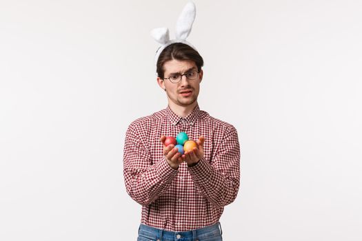 Traditions, religious holidays, celebration concept. Skeptical unsure cute guy dont like celebrating Easter day, look doubtful at painted eggs, feeling stupid in rabbit ears, stand white background.