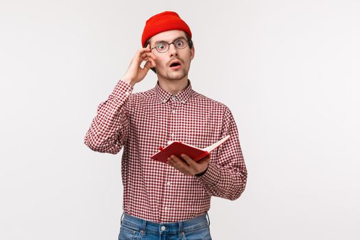 Waist-up portrait geeky and creative guy in red beanie, glasses have stunning great idea, touch temple and look inspired while taking notes in notebook, creating equation, stand white background.