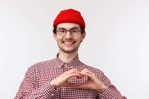 Close-up portrait cute bearded caucasian male in glasses and red beanie, smiling pleased, showing heart gesture, express love or sympathy, love somone, standing white background upbeat.