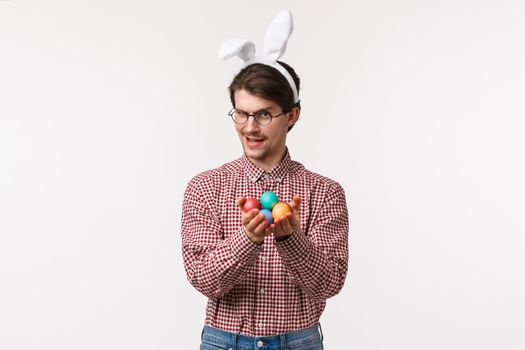 Traditions, religious holidays, celebration concept. Sassy and sly handsome caucasian male in glasses with rabbit ears, giving painted eggs and looking cunning camera, hinting, white background.